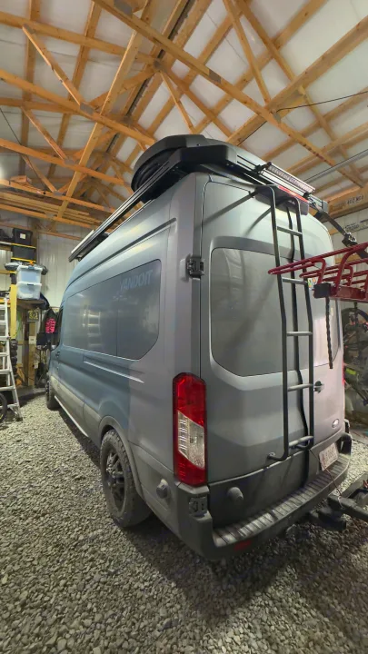 Gray Vandoit camper van (an rv rental option) with roof box, rear ladder, and bike rack, parked on gravel indoors.. Ford Custom Transit 2020