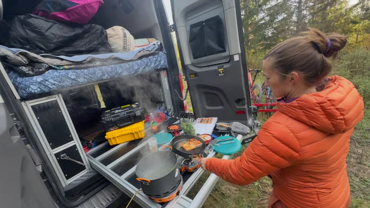 Cooking a fresh meal outside a camper van, possibly an rv rental. A person fries salmon and boils vegetables on portable stoves in a forest setting.. Ford Custom Transit 2020