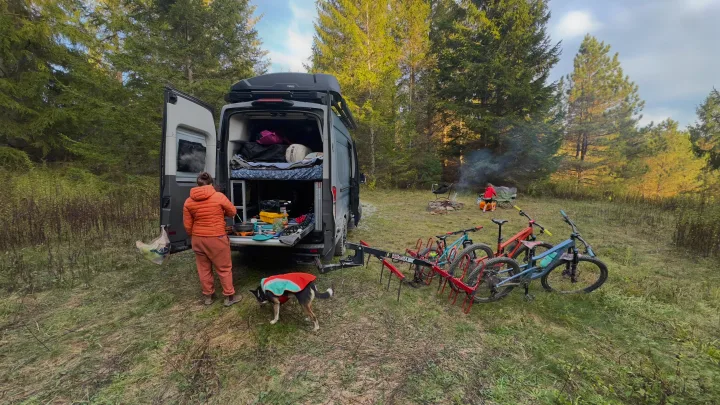 An RV rental provides a cozy camp setup: Cooking by the van, bikes, dog, and a child by the campfire.. Ford Custom Transit 2020