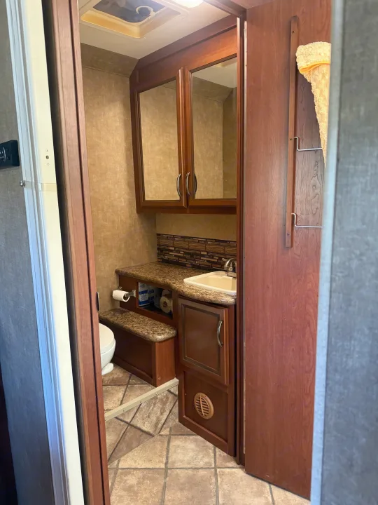 An RV rental bathroom: white toilet, brown wood vanity with white sink, mirror cabinet, and tiled floor. Earthy tones throughout.. Thor Chateau 2014