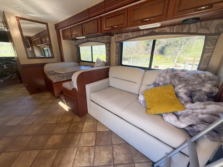 Bright RV rental interior: a light sofa with a plush blanket & yellow pillow, a dining booth, wooden cabinets, and windows revealing nature.. Thor Chateau 2014
