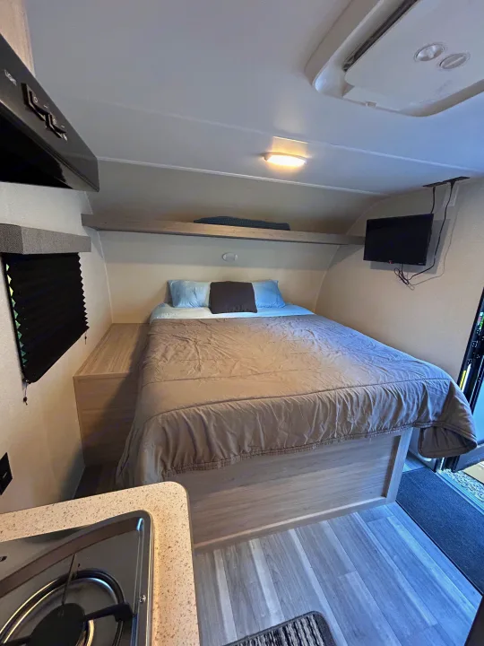 Cozy RV rental bedroom with a comfortable bed, overhead shelf, wall-mounted TV, and a partial view of the kitchen area.. Heartland RVs Pioneer 2025