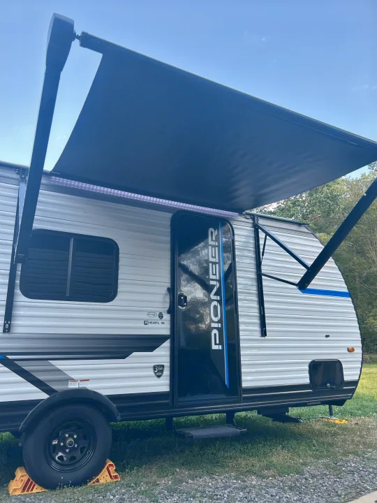 RV rental: A white Pioneer travel trailer with its extended black awning, glowing LED lights, and wheels chocked, parked on grass.. Heartland RVs Pioneer 2025