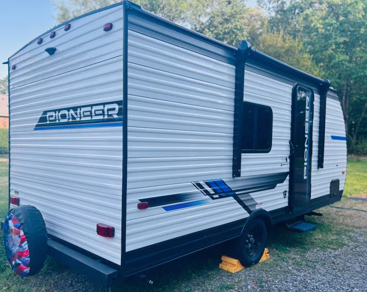 This white Pioneer travel trailer rv rental, with blue/black graphics and a patriotic spare tire, is parked on grass and gravel.. Heartland RVs Pioneer 2025