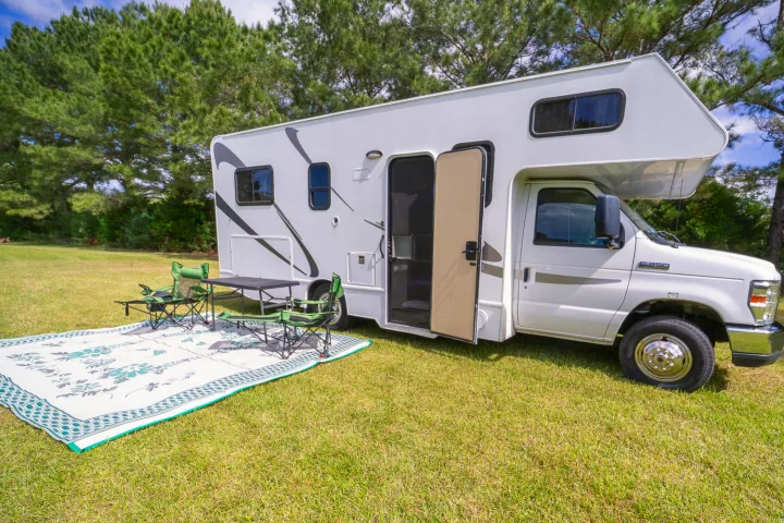 Class C RV rental parked on green grass with open door. Outdoor mat, table, and green chairs set up for camping.. Thor Majestic 2018