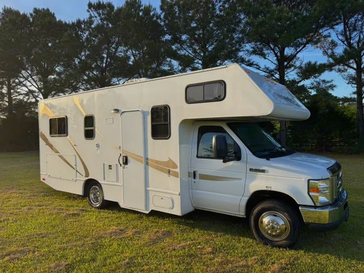 White Class C RV rental motorhome on green grass, backed by pine trees against a blue sky, perfect for travel.. Thor Majestic 2018
