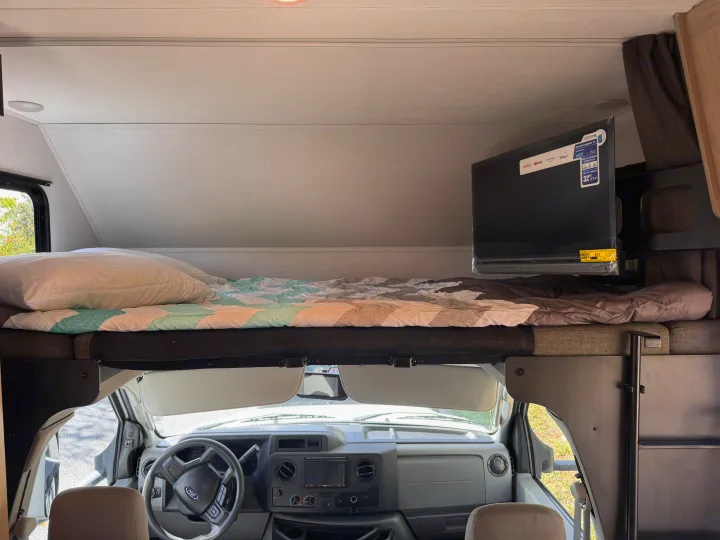 Inside an RV rental: a snug cab-over bed, Smart TV, and the Ford E-Series driver's area with dashboard and steering wheel.. Forest River Sunseeker LE 2025