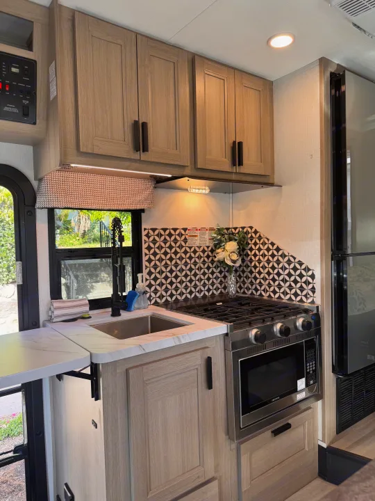 Modern RV rental kitchen with light wood cabinets, white countertops, sink, stove, and a distinctive black and white tiled backsplash.. Forest River Sunseeker LE 2025