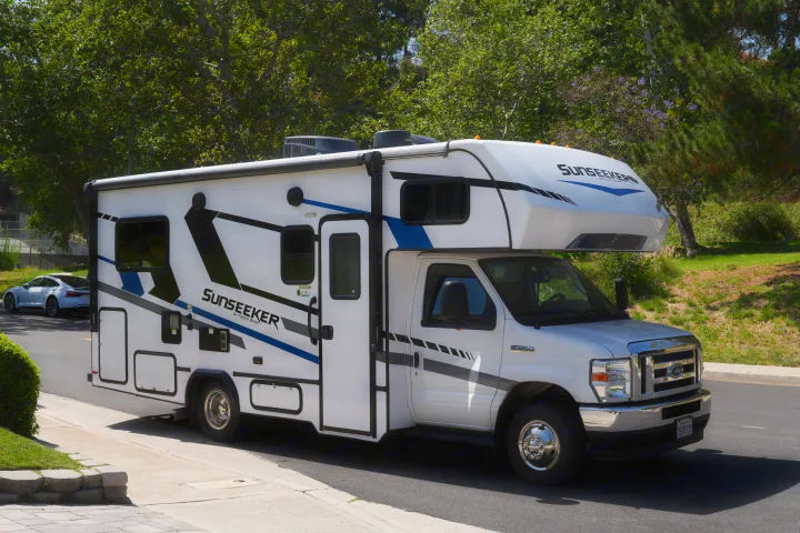 This white Sunseeker Class C RV rental, featuring blue and black graphics, is parked on a sunny street surrounded by trees.. Forest River Sunseeker LE 2025