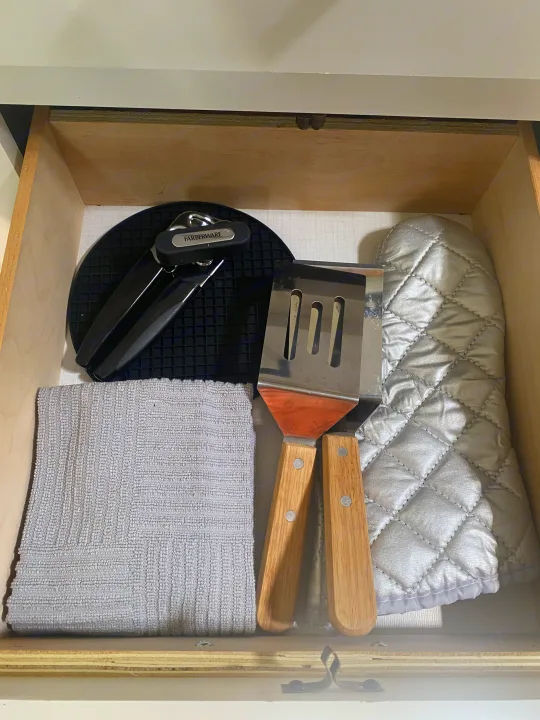 Cooking tools & linens in this RV rental drawer: can opener, spatula, oven mitt, silicone hot pad, and grey dish towel.. Keystone RV Hideout 2022