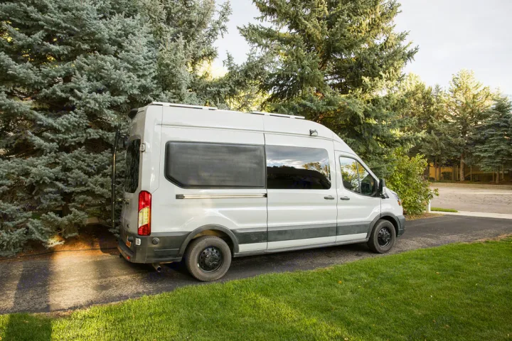 White camper van (RV rental) parked on driveway amidst tall trees & green grass.. Ford Transit 2022