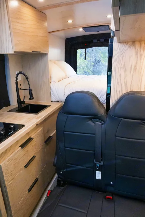 Modern RV rental interior: compact kitchen with sink, bed, and passenger seats visible. Light wood tones create a bright, functional space.. Ford Transit 2022