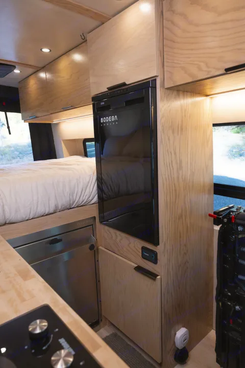 Experience an rv rental's cozy interior: light wood cabinetry, BODEGA COOLER fridge, bed with white linens, & cooktop.. Ford Transit 2022