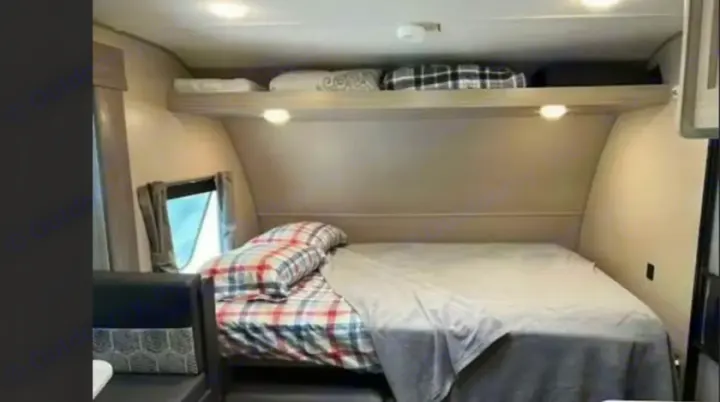 Inside an RV rental: a compact bedroom with a plaid bed, cozy grey blanket, overhead storage, and a window.. Coachmen Catalina Summit 2022