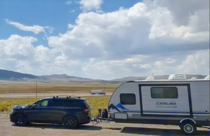 Black SUV tows a white Catalina rv rental trailer, parked by a highway with mountains and big clouds in the distance.. Coachmen Catalina Summit 2022