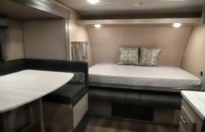 This cozy rv rental interior features a comfortable bed with decorative pillows, overhead lights, and a dining booth with a white table.. Coachmen Catalina Summit 2022