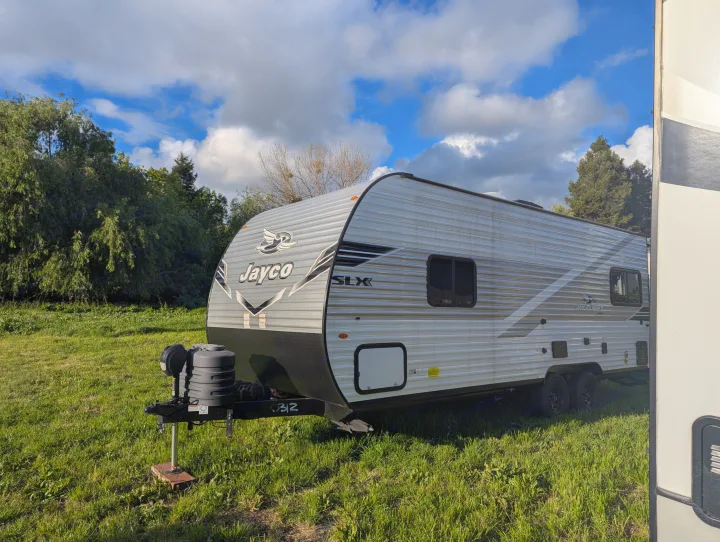 This Jayco SLX travel trailer RV rental is parked in a green field under a cloudy sky, surrounded by trees.. Jayco Jay Flight SLX 2025