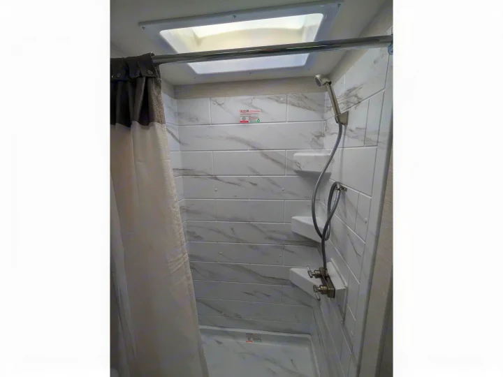 Modern RV rental shower featuring white marble-look tiles, a handheld shower, and an overhead skylight for bright, comfortable bathing.. Thor Chateau 2025