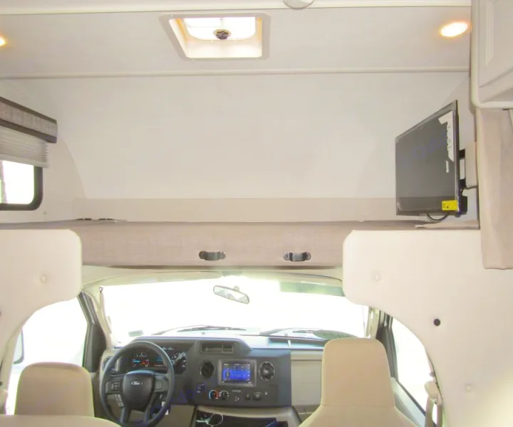 Interior of an RV rental: front cab with dashboard & steering wheel, and a cozy cab-over bunk bed above, with a TV.. Thor Chateau 2025