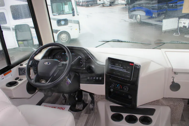 Inside a new **RV rental** cockpit: driver's seat, steering wheel, dashboard, and infotainment screen, with a rainy view outside.. Thor Motor Coach INDIGO - CC35 2024