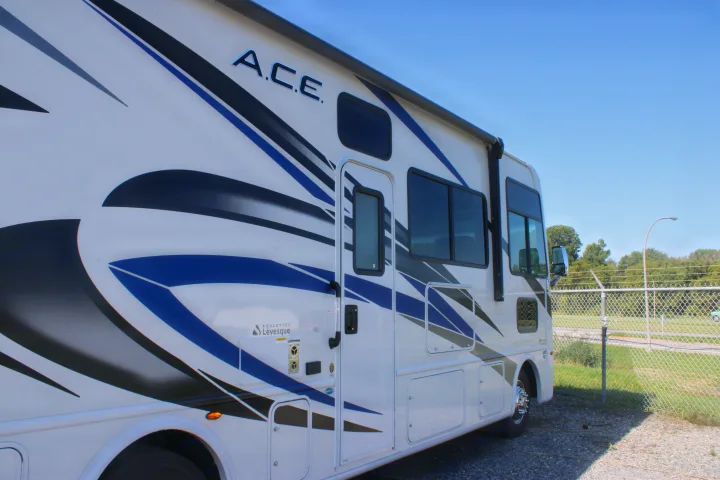 Sleek white A.C.E. RV rental, a Class A motorhome with blue & black graphics, parked on gravel on a sunny day.. Thor Motor Coach Ace 2024