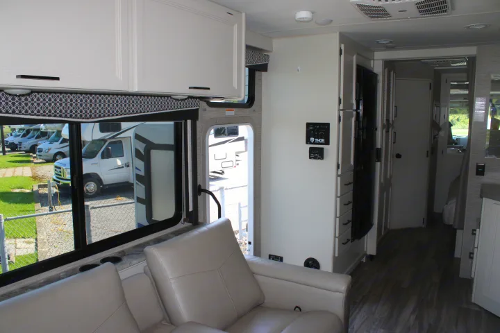 Interior of a modern RV rental, showing the living area with sofa, overhead cabinets, control panel, and hallway.. Thor Motor Coach Ace 2024
