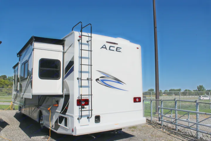 White A.C.E. RV rental with extended slide-out, ladder, and taillights, parked on gravel next to a chain-link fence.. Thor Motor Coach Ace 2024