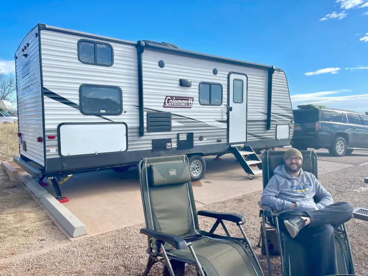Smiling man relaxing in a chair next to his Coleman RV rental travel trailer, with an SUV parked nearby.. Dutchmen Coleman Lantern 2020