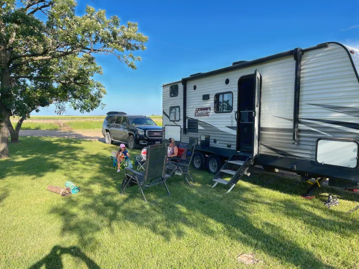 People relax by their RV rental. A Coleman travel trailer and GMC SUV are parked under a tree in a sunny, grassy field.. Dutchmen Coleman Lantern 2020