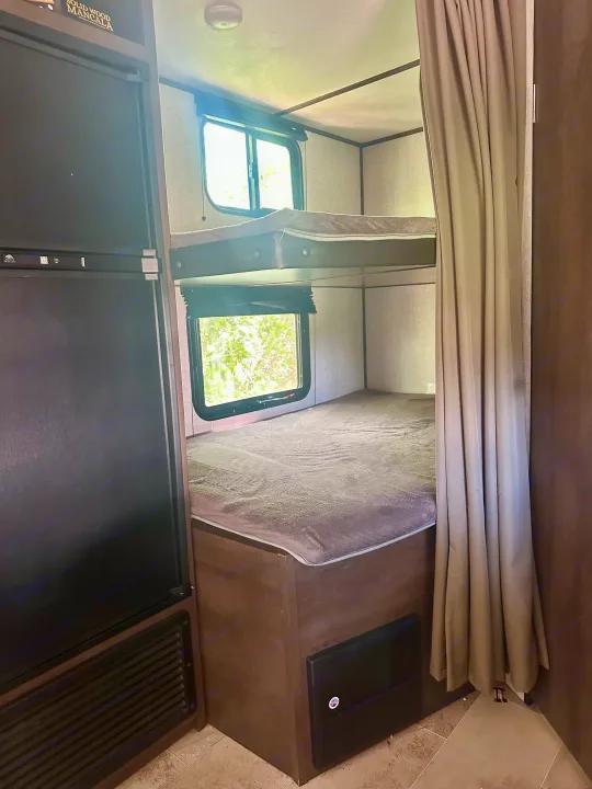 Inside an RV rental: two comfy bunk beds with windows, next to a refrigerator and a privacy curtain.. Dutchmen Coleman Lantern 2020