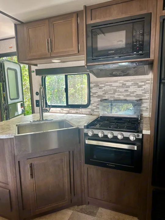 RV rental kitchen with a stainless sink, Furrion appliances (microwave, oven, range hood), wood cabinets, and a tiled backsplash.. Dutchmen Coleman Lantern 2020