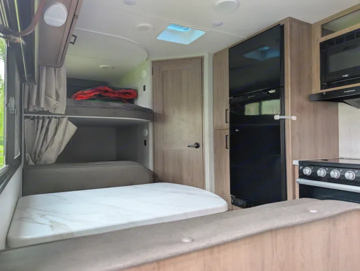 Explore this RV rental's interior: bunk beds with curtains, dinette table, and a compact kitchen area with fridge and stove.. Grand Design Imagine XLS 2023