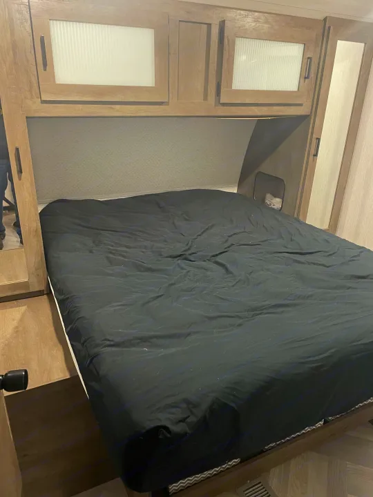 An RV rental's bedroom shows a bed with dark bedding, overhead wooden cabinets, and a side storage cubby. Modern camper interior.. Forest River Salem 2022