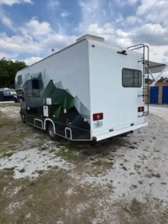 This RV rental features a modern green mountain side design, rear ladder, and extended awning, parked on a sunny day for travel adventure.. Thor Quantum LC 2022