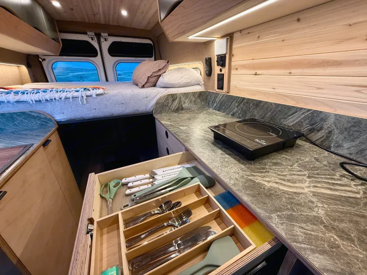 Cozy RV rental interior with a comfortable bed, a well-equipped kitchen counter featuring a cooktop, and an organized utensil drawer.. Ram Promaster 2015