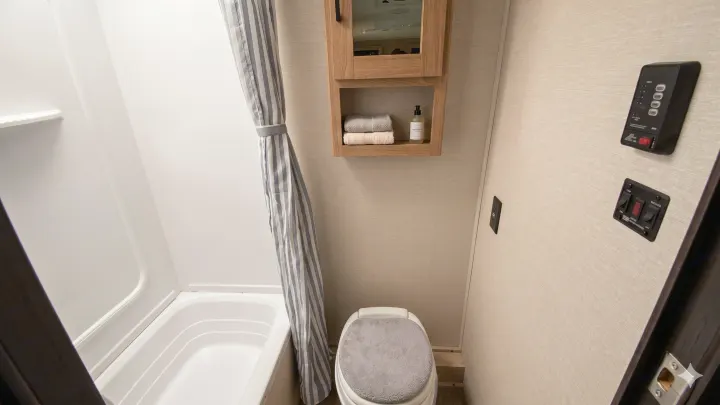 Compact RV rental bathroom: shower/tub, toilet with gray cover, wooden cabinet, and controls on a neutral wall.. Coachmen Apex Nano 2023