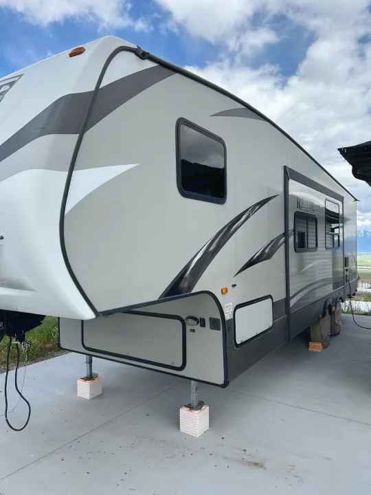 Modern grey and white fifth-wheel RV rental parked on concrete with leveling jacks. Mountains and sky are visible in the background.. Crossroads RV Rezerve 2016