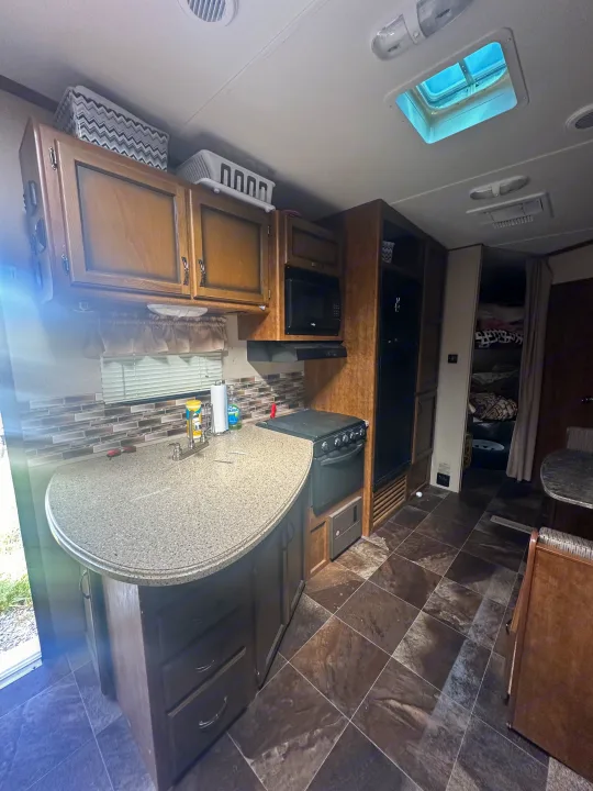 RV rental kitchen: light wood cabinets, speckled countertop with sink, stove, microwave, and dark tiled floor with skylight.. Crossroads RV Rezerve 2016