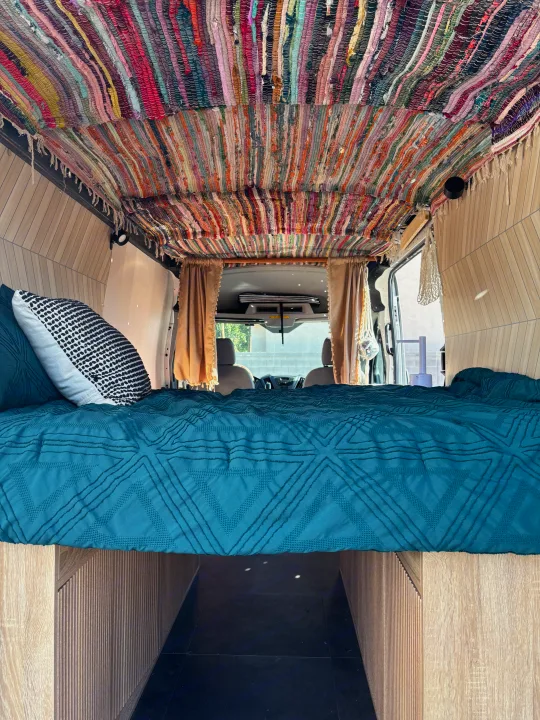 Colorful RV rental bedroom with a vibrant rag rug ceiling, warm wood paneling, and a cozy teal bed.. Ford E350 Box Van 2015