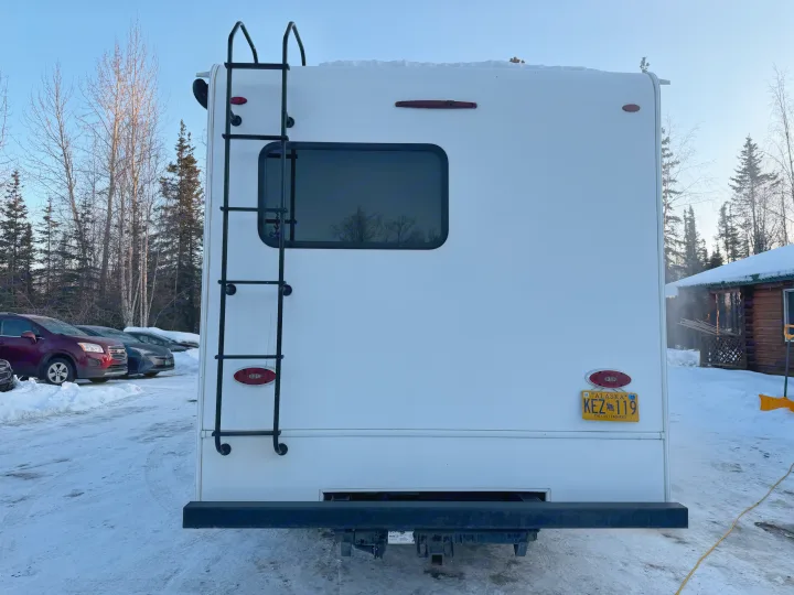 Rear view of a white RV rental in snowy Alaska, showing a ladder, window, and license plate KEZ 119 amidst winter trees.. Forester Forester Motorhome 2020