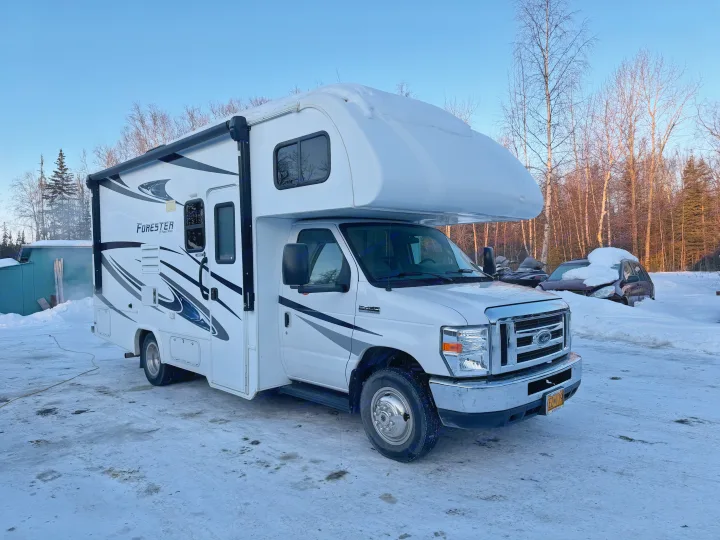 A white RV rental motorhome, snow-covered on top, is parked on icy ground in a winter setting with bare trees and a clear blue sky.. Forester Forester Motorhome 2020