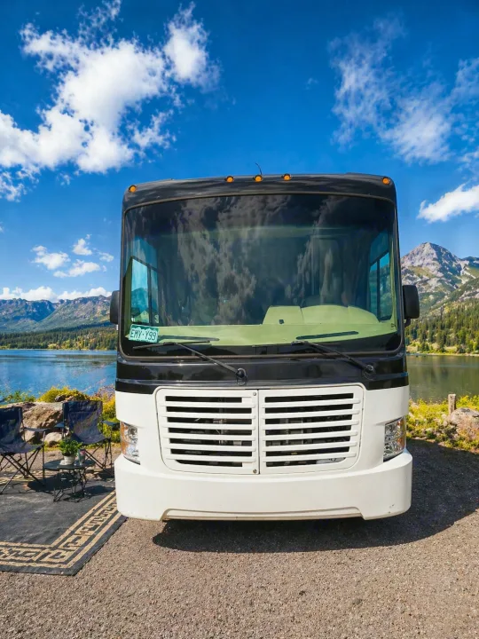 An RV rental ready for adventure. Front view of a motorhome by a lake, mountains, and a camp setup with chairs and a mat.. Coachmen Pursuit 2014