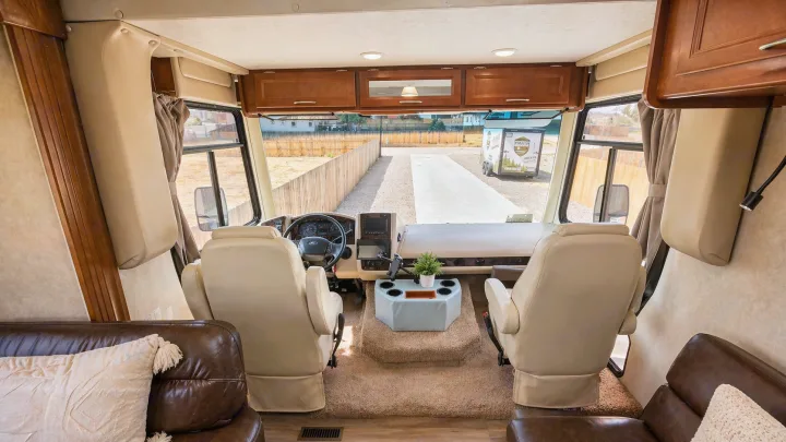 Spacious RV rental cockpit: cream seats, detailed dashboard, overhead storage, and a clear view of the fenced property ahead.. Coachmen Pursuit 2014