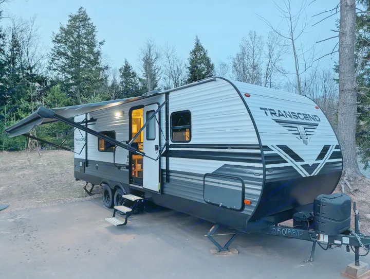 Enjoy your rv rental: A Transcend travel trailer with awning out & lights on, nestled among trees at twilight.. Grand Design Transcend 2020