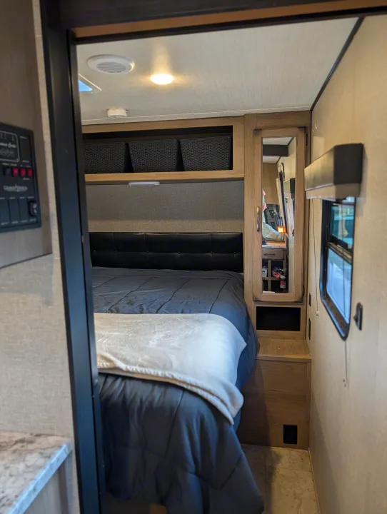 Cozy RV rental bedroom features a comfy bed with a dark comforter, upholstered headboard, overhead storage baskets, and a mirrored cabinet.. Grand Design Transcend 2020
