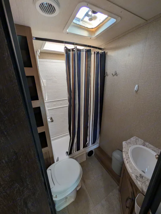 RV rental bathroom: Compact space with toilet, shower, sink, and vanity. Features a striped curtain, ceiling vent, and skylight.. Grand Design Transcend 2020