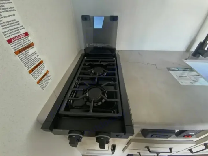 This rv rental kitchen features a black two-burner gas stovetop with a clear cover, light countertop, and safety warnings on the wall.. Forest River Puma 2025