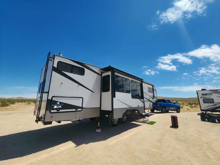 Large white Alliance RV rental with awning extended, parked in a desert landscape under a blue sky, with a blue truck behind it.. Alliance RV Avenue 2022