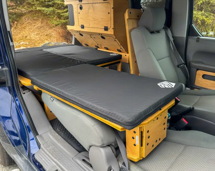 RV rental: Interior of a camper van with a black modular bed, wooden storage, and a front passenger seat, ready for road trips.. Honda Element 2010
