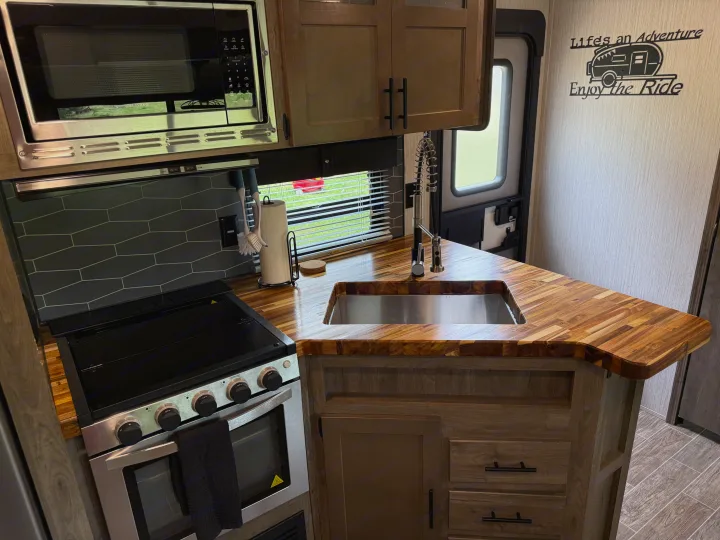 Interior of an rv rental kitchen with a microwave, stove/oven, butcher block counters, sink, and "Life's an Adventure" decal.. Forest River Palomino Puma 2021